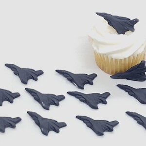 Puede incluir: Una colección de once decoraciones para pasteles con forma de avión de combate en color gris oscuro. Diez de las figuras están dispuestas en filas, mientras que una se coloca sobre un cupcake con glaseado blanco. Los jets tienen alas y cola.