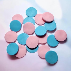 May include: A collection of round, flat objects in shades of pink and teal. The objects are scattered across a white and pink gradient background, creating a visually appealing arrangement. The objects appear to be made of a soft material.