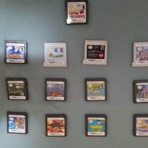 May include: A collection of Nintendo DS game cartridges arranged on a light blue surface. The cartridges feature various game titles, with colorful artwork and text labels. The games are in their original cases.
