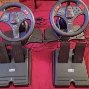 Könnte beinhalten: Zwei schwarze V3-Rennlenkräder mit roten Knöpfen und angeschlossenen Pedalen. Die Lenkräder haben ein PlayStation-Logo. Die Pedale befinden sich auf einer schwarzen Basis, vor rotem Hintergrund.