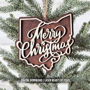 May include: A wooden Christmas ornament in the shape of Ohio, with the words "Merry Christmas" in elegant script. The ornament is brown with a lighter wood outline and text, hanging from a natural twine string. The ornament is displayed on a green Christmas tree.