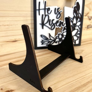 May include: A black wooden easel stand with two angled legs and a flat top. The stand is designed to hold a decorative sign or plaque.