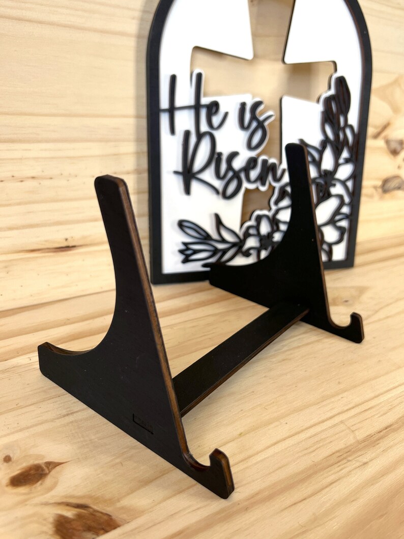 May include: A black wooden easel stand with two angled legs and a flat top. The stand is designed to hold a decorative sign or plaque.