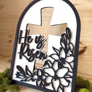 May include: White wooden sign with a black cross and floral design. The sign reads "He is Risen".