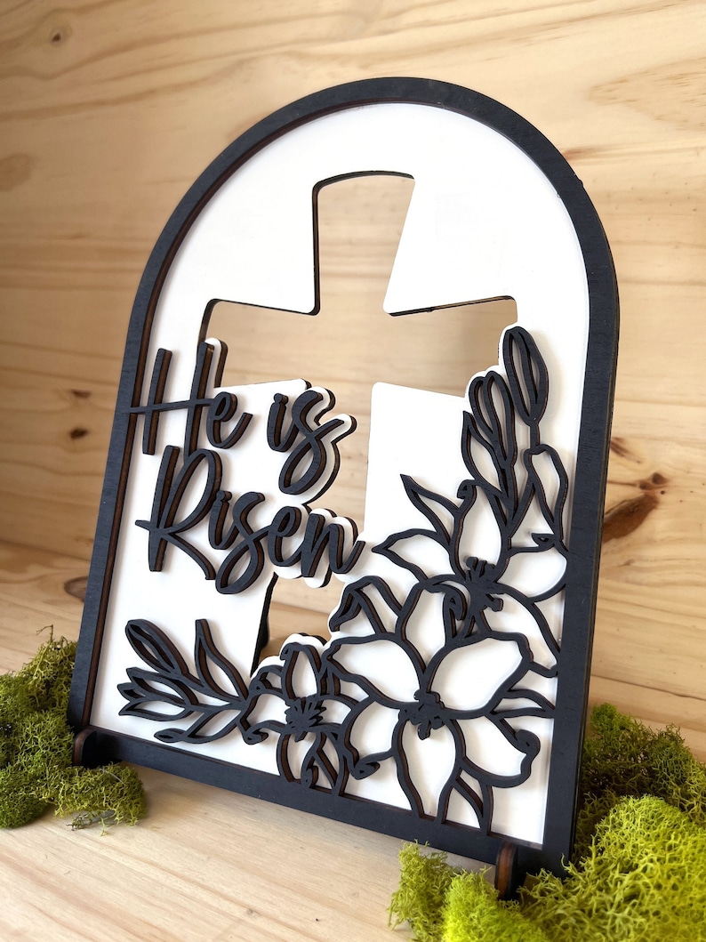 May include: White wooden sign with a black cross and floral design. The sign reads "He is Risen".