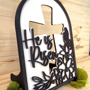 May include: A white wooden cross with the words "He is Risen" in black lettering, surrounded by a floral design. The cross is mounted on a black wooden stand.