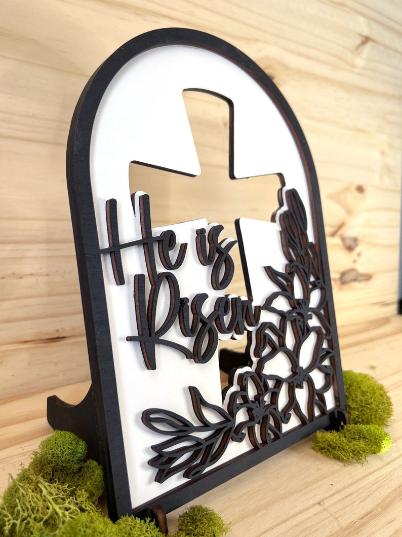 May include: A white wooden cross with the words "He is Risen" in black lettering, surrounded by a floral design. The cross is mounted on a black wooden stand.