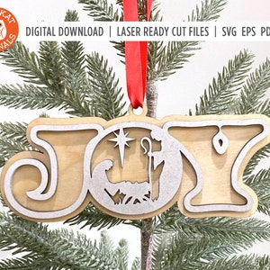 May include: A wooden Christmas ornament with the word "JOY" in a stylized font. The "O" is a circle with a silver glitter finish and a nativity scene inside. The ornament is hanging on a green pine branch.