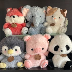 May include: A selection of soft plush animal toys. The collection includes a red fox, a grey elephant, a white and brown cat, a grey penguin, a pink pig, and a black and white panda. Each toy has a round tag.