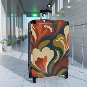 Puede incluir: Una maleta con ruedas con un diseño floral retro en tonos naranja, crema y verde azulado. Las palabras "Peace", "Love" y "Happiness" están impresas en la parte delantera. La maleta tiene un asa retráctil y cuatro ruedas.