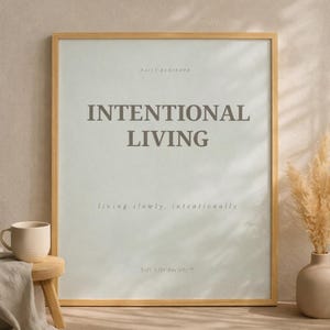 Minimalist printable wall art poster featuring the affirmation “INTENTIONAL LIVING” in elegant modern serif typography on a soft neutral sage green background. Poster displayed in a light natural wood frame styled in a calm Scandinavian interior with warm natural sunlight and soft shadows. Feminine quiet luxury home decor aesthetic.