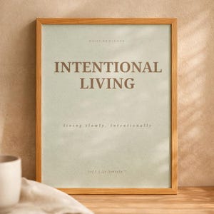 Close-up detail of minimalist affirmation poster reading “INTENTIONAL LIVING” showing elegant serif typography, subtle paper texture, and natural wood frame in warm editorial sunlight. Soft luxury printable wall art styling