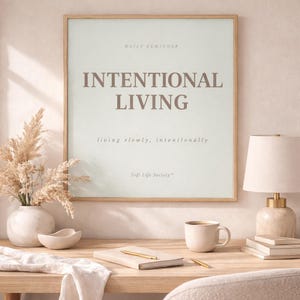 Minimalist framed wall art print featuring the quote “INTENTIONAL LIVING” displayed above a clean light wood desk with neutral stationery, journal, coffee mug, and warm natural lighting. Calm feminine workspace decor aesthetic.