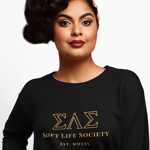 Könnte beinhalten: Schwarzes Sweatshirt mit goldfarbenem Schriftzug „ΣΛΣ SOFT LIFE SOCIETY EST. MMXXV“. Das Model hat dunkles Haar und trägt dunklen Lippenstift. Das Sweatshirt ist ein Rundhals-Stil.