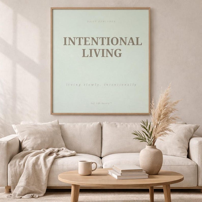 Framed printable affirmation poster reading “INTENTIONAL LVING” styled in a modern minimalist home setting with neutral decor, soft linen textures, ceramic mug, pampas grass, and warm morning light creating a peaceful soft life atmosphere.