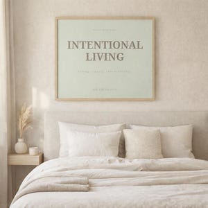 Minimalist printable affirmation wall art poster reading “INTENTIONAL LIVING” displayed above a soft neutral bed in a calm Scandinavian bedroom interior. Light natural wood frame, warm morning sunlight, linen bedding, ceramic decor accents, and peaceful feminine soft life aesthetic.
