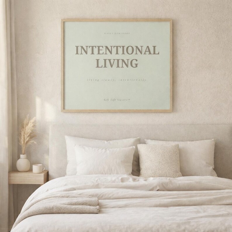 Minimalist printable affirmation wall art poster reading “INTENTIONAL LIVING” displayed above a soft neutral bed in a calm Scandinavian bedroom interior. Light natural wood frame, warm morning sunlight, linen bedding, ceramic decor accents, and peaceful feminine soft life aesthetic.