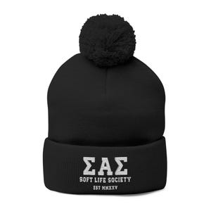 Può includere: Berretto nero lavorato a maglia con pompon. Il berretto presenta un testo ricamato bianco: "ΣΑΣ SOFT LIFE SOCIETY EST MMXXV". Un caldo cappello invernale.