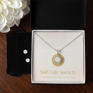May include: A silver necklace with a round, clear stone pendant, presented in a white box with the text "Soft Life Society". The box also contains matching stud earrings and a black velvet pouch. A white flower is in the background.