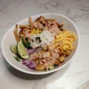 Può includere: Una ciotola bianca piena di un'insalata colorata con pollo, mais, formaggio, strisce di tortilla, riso, cipolla rossa e spicchi di lime. L'insalata è ricoperta da una salsa rosa cremosa.