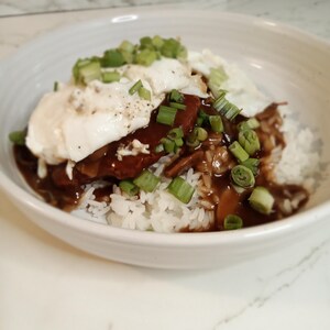 May include: A white bowl filled with a Hawaiian dish called Loco Moco. The dish consists of white rice topped with a hamburger patty, gravy, a fried egg, and green onions.