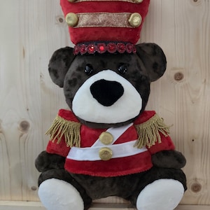 May include: A plush bear wearing a red soldier uniform with gold fringe epaulets, a white cross-body strap with gold buttons, and a matching red hat featuring gold trim, gold buttons, and a band of red sequins around the base of the hat.