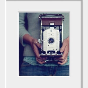 Może przedstawiać: Antyczny aparat Polaroid Land w kolorze srebrnym z brązowym sk&oacute;rzanym paskiem. Aparat jest trzymany przez osobę ubraną w niebieski sweter i dżinsy.