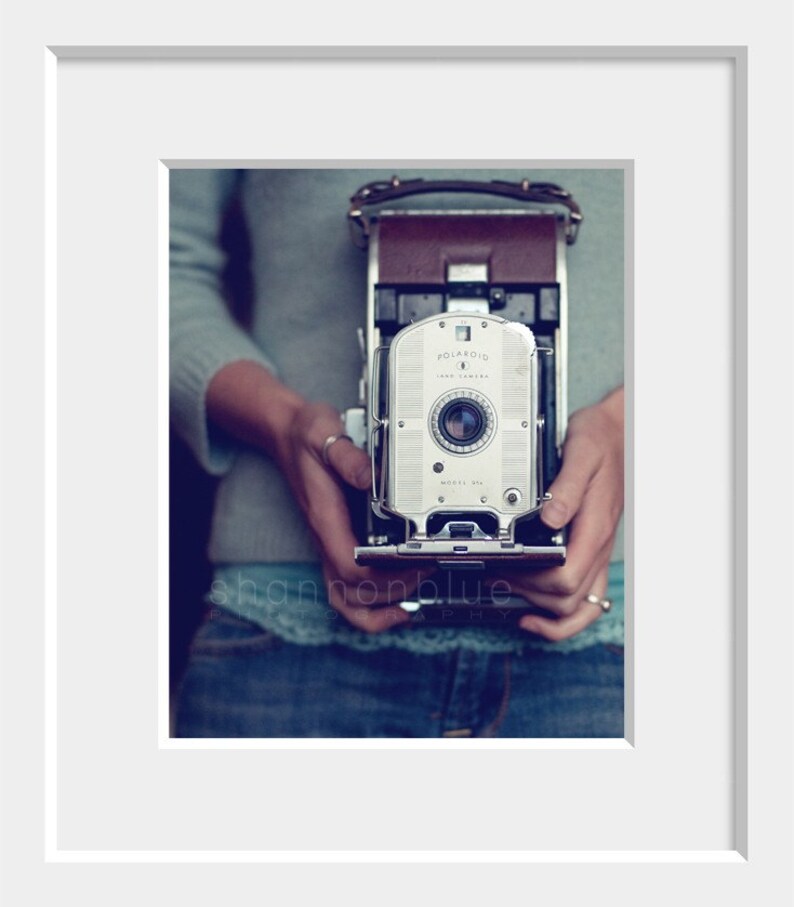 Może przedstawiać: Antyczny aparat Polaroid Land w kolorze srebrnym z brązowym sk&oacute;rzanym paskiem. Aparat jest trzymany przez osobę ubraną w niebieski sweter i dżinsy.