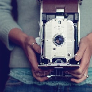 Może przedstawiać: Srebrny aparat Polaroid Land Camera, model 35, z brązowym paskiem sk&oacute;rzanym. Aparat jest trzymany przez osobę ubraną w niebieski sweter i dżinsy.