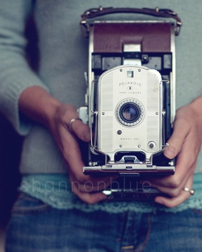 Może przedstawiać: Srebrny aparat Polaroid Land Camera, model 35, z brązowym paskiem sk&oacute;rzanym. Aparat jest trzymany przez osobę ubraną w niebieski sweter i dżinsy.
