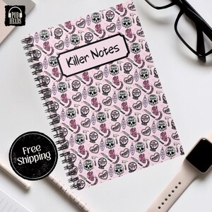 May include: Spiral-bound notebook with a pink cover featuring a repeating pattern of skulls, hearts, knives, and axes. The notebook has the words "Killer Notes" in a white rectangle. The notebook is on a white surface.