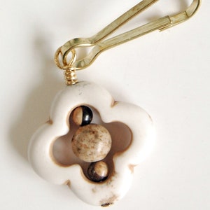 May include: A cream-colored clover-shaped charm with a gold-tone clasp. The charm features three spherical beads in shades of brown and black. The clasp is attached with a gold-tone wire.