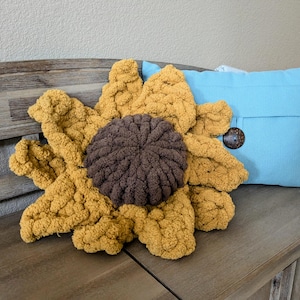 Op de afbeelding: Een handgemaakt, oversized zonnebloemkussen met een bruin midden en goudgele bloemblaadjes. Een lichtblauw kussen met een grote houten knoop staat op de achtergrond. De kussens liggen op een houten bank.