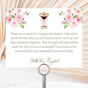 Può includere: Un biglietto bianco con testo e decorazioni floreali, tenuto da un supporto in metallo argentato. Il biglietto presenta un'illustrazione ad acquerello di un cocktail e rose rosa. Il testo recita: "Grazie mille per essere venuti alla mia festa!" e "Con amore, Crystal."