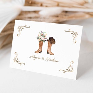 May include: A white card with a gold border and a floral design. The card features a pair of brown cowboy boots with a bouquet of flowers and a cowboy hat. The text "Alyssa & Nathan" is printed below the boots.