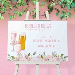 May include: A white sign with pink text and floral accents announcing a "Bubbles & Brews" baby shower for Danielle & Michael. The sign features illustrations of a champagne flute, a beer glass, and a baby bottle.