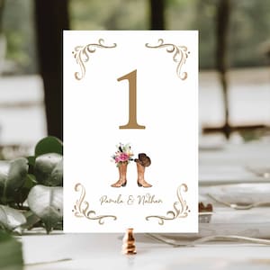 May include: A white table number card with gold accents, featuring the number 1 in gold. Below the number are illustrations of cowboy boots, a hat, and flowers. The names "Pamela & Nathan" are printed below the illustrations.