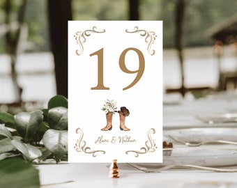 May include: A white table number card with the number 19 in gold. The card features a watercolor illustration of cowboy boots and a hat, with floral accents. The names "Alani & Nathan" are printed below the illustration. Decorative gold flourishes frame the top and bottom.