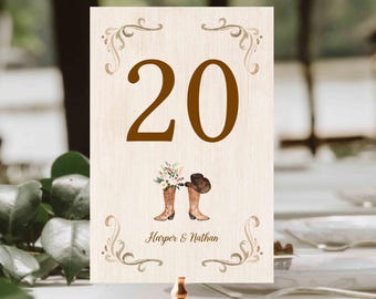 May include: A rustic wedding table number card featuring the number 20 in brown. The card includes a watercolor illustration of cowboy boots with flowers and a hat. The names "Harper & Nathan" are printed below the illustration.