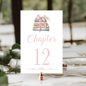 Puede incluir: Una tarjeta blanca con el número de mesa con una ilustración de acuarela de una pila de libros atados con una cinta rosa y flores. La tarjeta presenta la palabra "Chapter" y el número "12" en escritura rosa. Un soporte dorado decorativo sostiene la tarjeta.