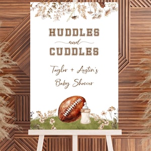 Op de afbeelding: Een wit bord op een ezel met de tekst "Huddles and Cuddles" en "Taylor + Austin's Baby Shower". Het bord toont een voetbal en een babyfles met bloemaccenten. De achtergrond is een houten muur.