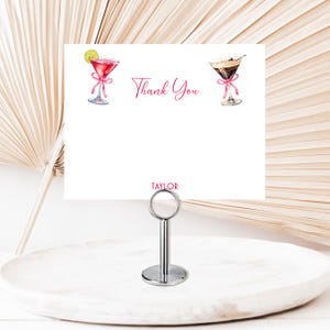 May include: A white card with the words "Thank You" in pink script, featuring watercolor illustrations of cocktails. The card is held by a silver metal stand, perfect for table settings or place cards.