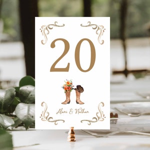 May include: White table number card with the number 20 in gold. The card features a watercolour illustration of cowboy boots and a hat, with the names Alani & Nathan below. Decorative gold flourishes frame the top and bottom.