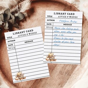 May include: Two white library cards with the text "LIBRARY CARD ADVICE & WISHES". One card has handwritten messages in blue ink. Both cards feature a small illustration of pumpkins and books. The cards are on a wooden tray.