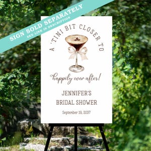 Puede incluir: Cartel blanco sobre un caballete negro con el texto "A 'Tini Bit Closer To" y "Happily ever after!" con una ilustraci&oacute;n de acuarela de un martini. El cartel es para la despedida de soltera de Jennifer, 19 de septiembre de 2037.