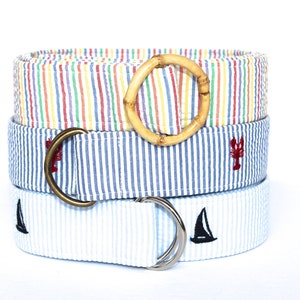 May include: Three fabric belts stacked on top of each other. The top belt has a bamboo buckle and is striped with red, yellow, blue, and green. The middle belt is blue and white striped with a red lobster. The bottom belt is light blue and white striped with a black sailboat.