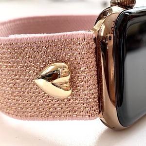 May include: A rose gold Apple Watch with a pink and gold glitter elastic band and a gold heart charm.
