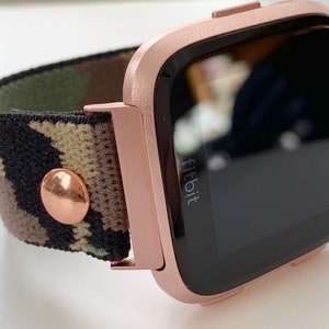 May include: Rose gold Fitbit smartwatch with a camouflage patterned band. The watch face is black and displays the word "fitbit".
