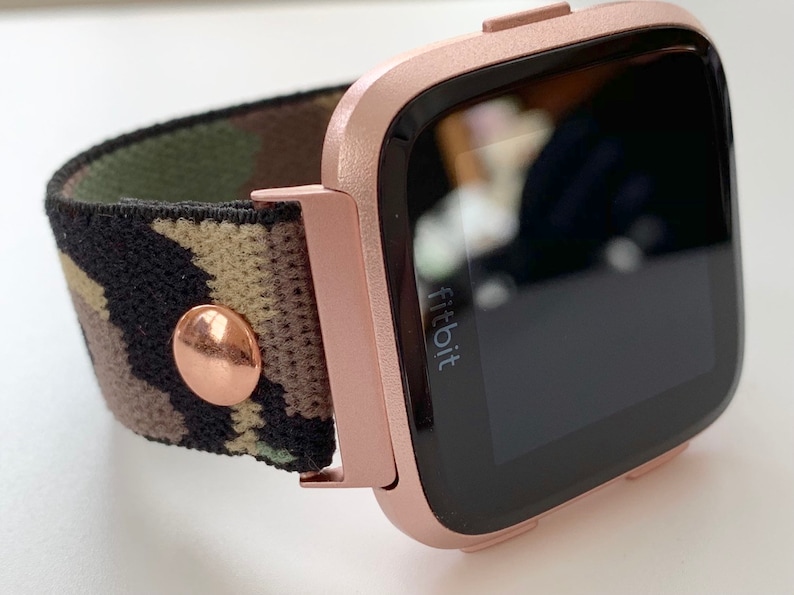 May include: Rose gold Fitbit smartwatch with a camouflage patterned band. The watch face is black and displays the word "fitbit".