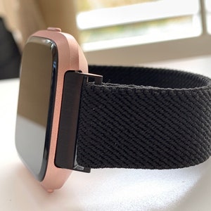 May include: A rose gold smartwatch with a black elastic band. The watch face is black and rectangular.